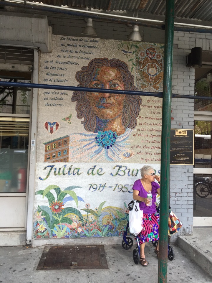 A woman sits in front of a mural by Manny Vega honoring Julia De Burgos.